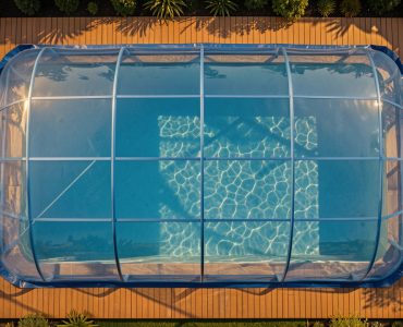 Piscine familiale rectangulaire vue en plongée, recouverte d’une bâche à bulles bleue, avec une terrasse en bois éclairée par la lumière dorée de fin de journée.